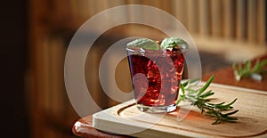 Red cocktail with ice, decorated with mint leaf on wooden cutting board with rosemary near, books on background, home interior