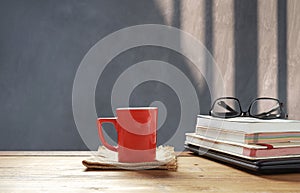 Red coffee cup and eyeglasses on stacked books, black laptop on wooden table in front of loft cement wall background