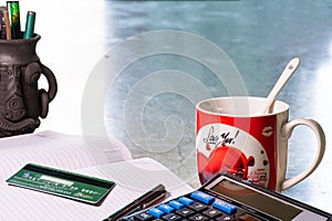 A red coffee mug with glass spoon and a diary, debit card, digital calculator, a pen, and soil pottery pen stand on the study desk