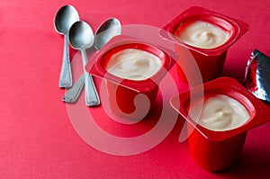 Red colour yogurt plastic pots with spoons and foil on red background - creamy fruit yoghurt in plastic cups with copy space