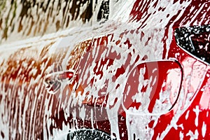 Red compact SUV car with sport and modern design washing with soap. Car covered with white foam. Car care service business concept