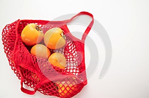 A red cotton string bag with persimmons and tangerines lies on a white background. The concept of saving the earth from plastic.
