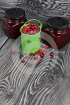 Red cranberries in a miniature green bucket. Stands on brushed boards. Nearby is jars of cranberry jam and some berries scattered