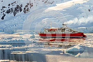 Red cruise steamboat drifting among the icebergs with huge rock and glacier in background, Neco bay, Antarctica