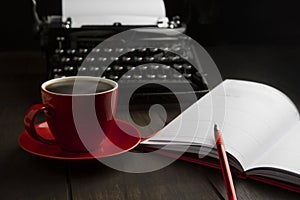 A red cup with coffee in close-up. An old vintage typewriter and an empty piece of paper. A notepad for writing and a