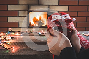 Red cup of coffee in female hand by the fireplace. Female relaxes by warmfire in christmas red socks. Christmas holiday.
