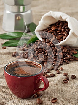 Red cup of coffee with smoke espresso and coffee beans in burlap sack on a burlap background. Close up, soft focus. Near coffee