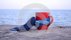 Red cup with hot coffee tea, tied with blue knitted scarf stands on stone