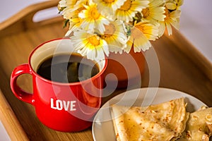 Red cup with the word `love` with hot coffee / tea, a bouquet of daisies and pancakes with honey on a wooden tray