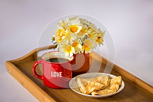 Red cup with the word `love` with hot coffee / tea, a bouquet of daisies and pancakes with honey on a wooden tray