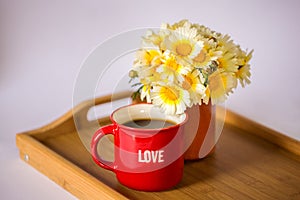 A red cup with the word `love` with hot coffee / tea and a bouquet of daisies on a wooden tray