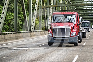 Red day cab big rig semi truck transporting liquid cargo in tank semi trailer driving on the highway road with truss bridge
