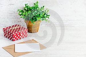 Red dotted gift box, empty card, kraft envelope and a green flower in a rustic ceramic pot. White wooden background, copy space.