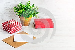 Red dotted gift box, empty card, kraft envelope, red book and a green flower in a rustic ceramic pot. White wooden background, cop