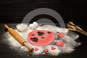 Red dough, cut out hearts, flour, eggs and rolling pin on a black background, ears of wheat, space for text