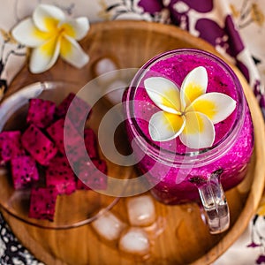 Red Dragon fruit juice in a glass, decorated with Plumeria flower, cut dragon fruit and ice cubes. Served on a round wooden tray o