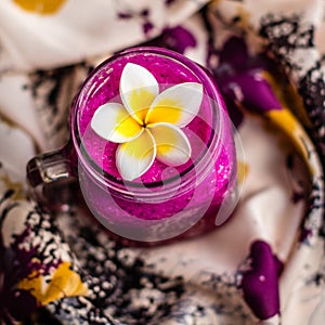 Red Dragon fruit juice in a glass, decorated with Plumeria flower over flowery fabric. Top view. Square image.