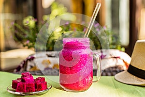 Red Dragon fruit juice in a glass with a glass straw, hat, cut dragon fruit on a glass plate. Flowers on a background. On a green