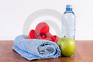 Red dumbbell on blue towel and green apple and water bottle on wooden floor