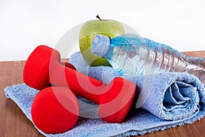 Red dumbbell, fresh water bottle with dew and green apple on blue towel