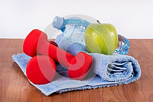 Red dumbbell, fresh water bottle with dew and green apple on blue towel on wooden floor