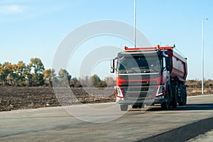 A red dump truck drives out of town on a flat asphalt road among fields against a blue sky on a sunny day