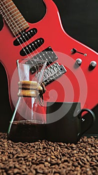 A red electric guitar on a black background, with a coffee brewing jug in front of it and a black coffee mug on coffee beans.