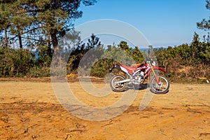 A red enduro Motorcycle parked on dirt road and stands. Trees and sea background