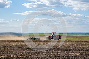 A red farm tractor in a cloud of dust cultivates the soil in the field with a cultivator after harvest. Summer sunny day