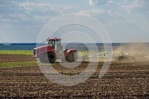 A red farm tractor in a cloud of dust cultivates the soil in the field with a cultivator after harvest. Summer sunny day