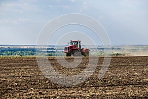 A red farm tractor in a cloud of dust cultivates the soil in the field with a cultivator after harvest. Summer sunny day