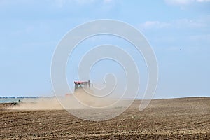 A red farm tractor in a cloud of dust cultivates the soil in the field with a cultivator after harvest. Summer sunny day