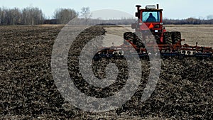 Red farm tractor pulling cultivator turning up black soil on field after harvest