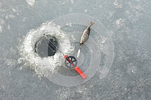 Red fishingrod and a silver lure next to a hole in the ice and a perch