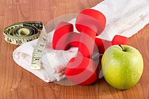 Red fitness dumbbell, measuring tape and green fresh apple with dew on white towel and wooden floor
