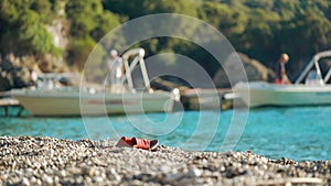 Red flip flop or sandal shoes on small pebbles beach, blurred calm turquoise sea with motor boats and trees behind. Afternoon