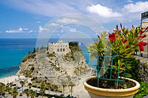 Red flowers in flowerpot with blurred Monastery Sanctuary church Santa Maria dell Isola on top of rock, Tyrrhenian Sea, blue sky