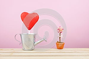 The red foiled chocolate heart stick with small silver watering can and mini fake flower in brown plant pot on wooden tray