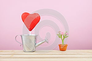 The red foiled chocolate heart stick with small silver watering can and mini fake flower in brown plant pot on wooden tray