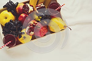 Red fresh juice with apples, pears, bananas, grapes and pomegranate fruits in white wooden tray on bed sheet.