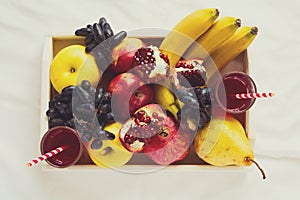 Red fresh juice with apples, pears, bananas, grapes and pomegranate fruits in white wooden tray on bed sheet. Top view. Healthy l