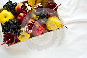 Red fresh juice with apples, pears, bananas, grapes and pomegranate fruits in white wooden tray on bed sheet. Top view. Healthy l