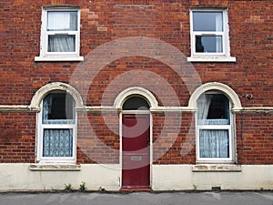 Red front door and windows of a typical old brick british terraced house with arched windows and net curtains