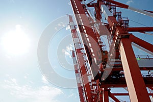 Red gantry cranes with white stripes in vertical position in container terminal with blue sky in background waiting for loading.