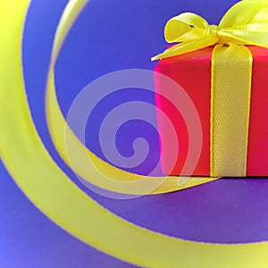 Red Gift box at the end of the spiral yellow ribbon, indigo background, square.