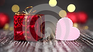Red gift box with golden knot and pink heart close-up. Bright decorative balls in the background. Christmas tree lights blink yell