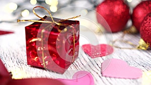 Red gift box with golden knot and pink hearts close-up. Bright decorative balls and ribbon. Christmas tree lights blink yellow