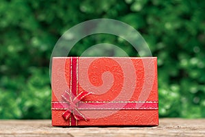 Red gift box with a red ribbon on wooden desk with a green leaves background. Celebration gift card