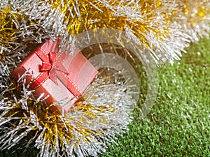 Red gift box with red ribbon bow and golden seam place on silver and gold rainbow glowing decoration background on green grass.