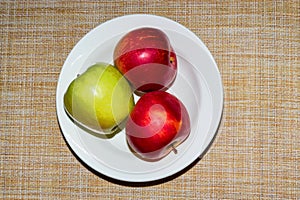 Red and green apple on a white plate and orange background viewed from above. Three ripe apples on a porcelain plate on a brown